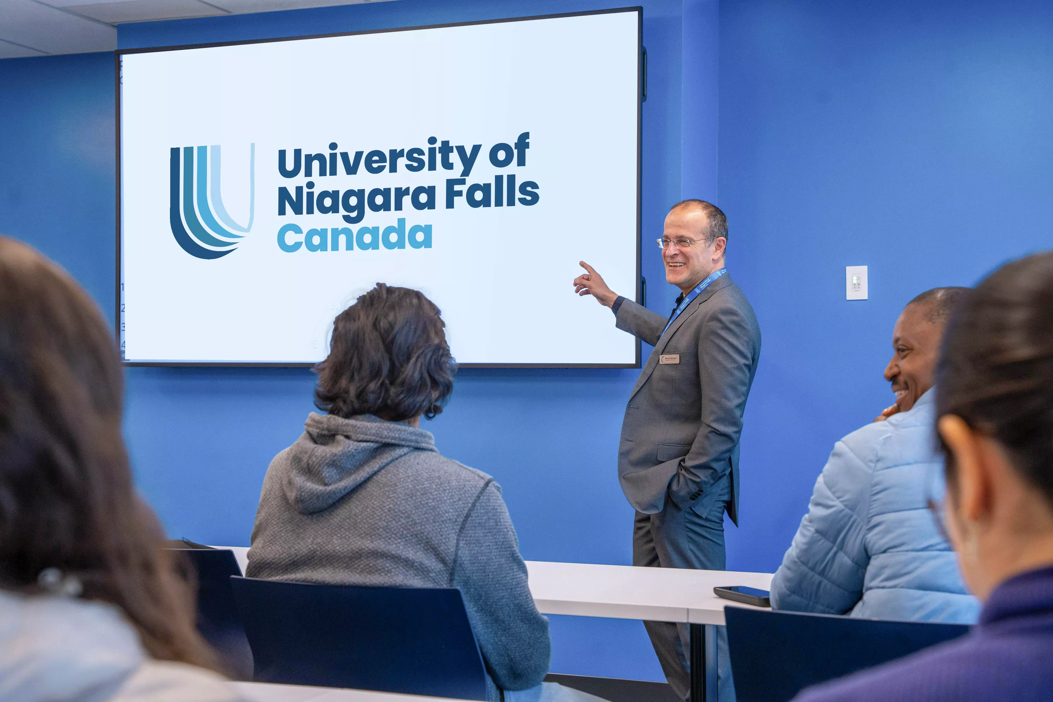 Male teacher in a suite standing in front of white display screen with &quot;University of Niagara Falls Canada&quot; in blue